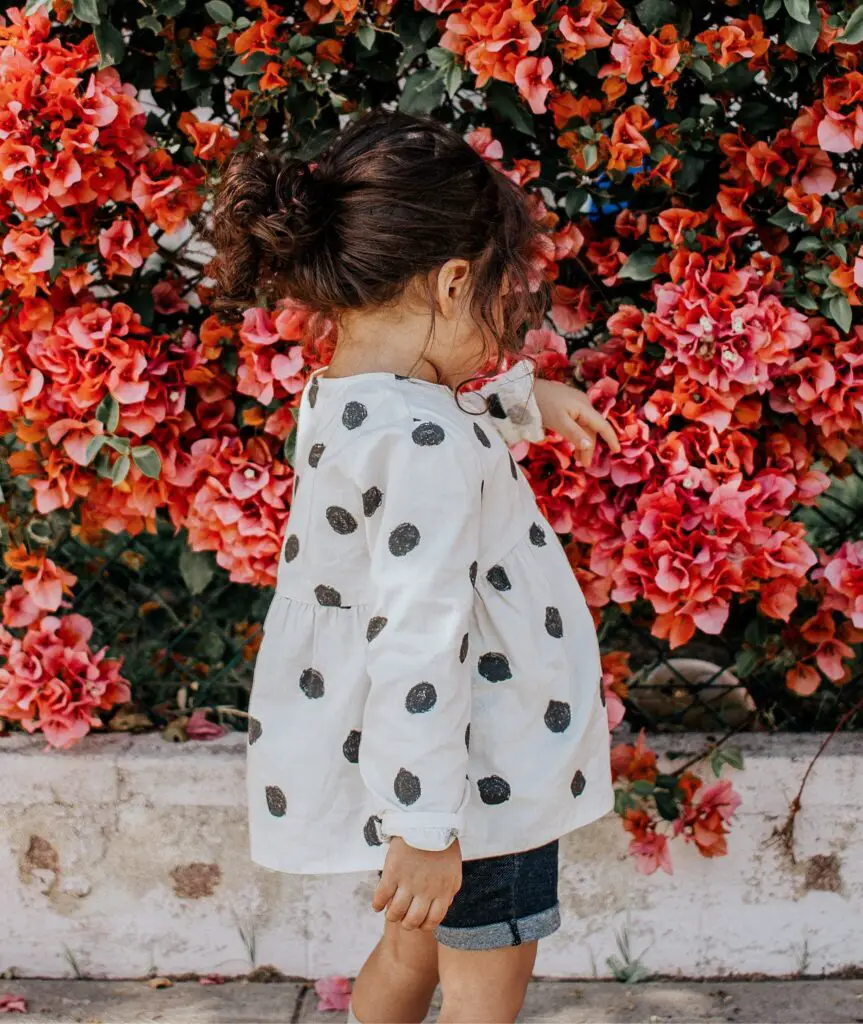 Little girl wearing a polka dot shirt and standing in front of red flowers. My kidizen review shares how you can make money reselling kid clothes. 